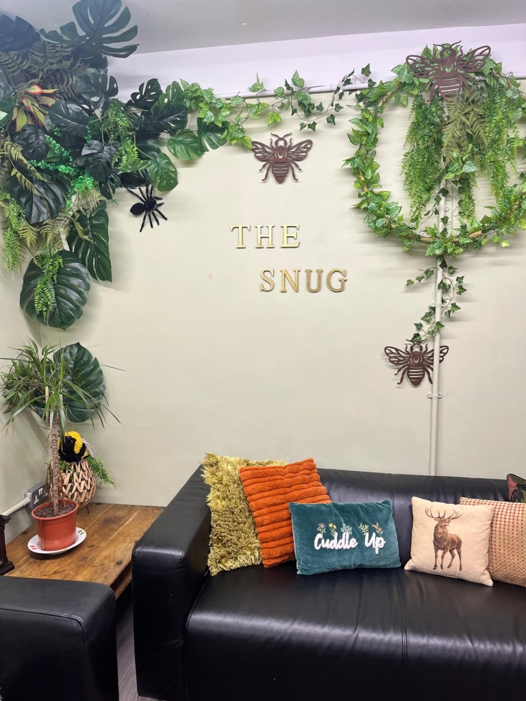The ARC 1 Snug feature wall, with gold THE SNUG lettering, metal bees and hanging plants above a sofa